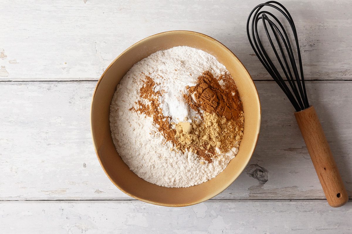 A beige bowl filled with flour, baking soda, ginger, and cinnamon on a white wooden surface. A whisk with a wooden handle rests beside the bowl.