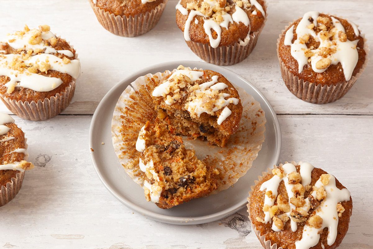 Seven muffins topped with white icing and nuts are on a white wooden surface. One muffin is cut in half on a gray plate, revealing a moist interior with visible nut pieces. The muffins have a light golden-brown color.