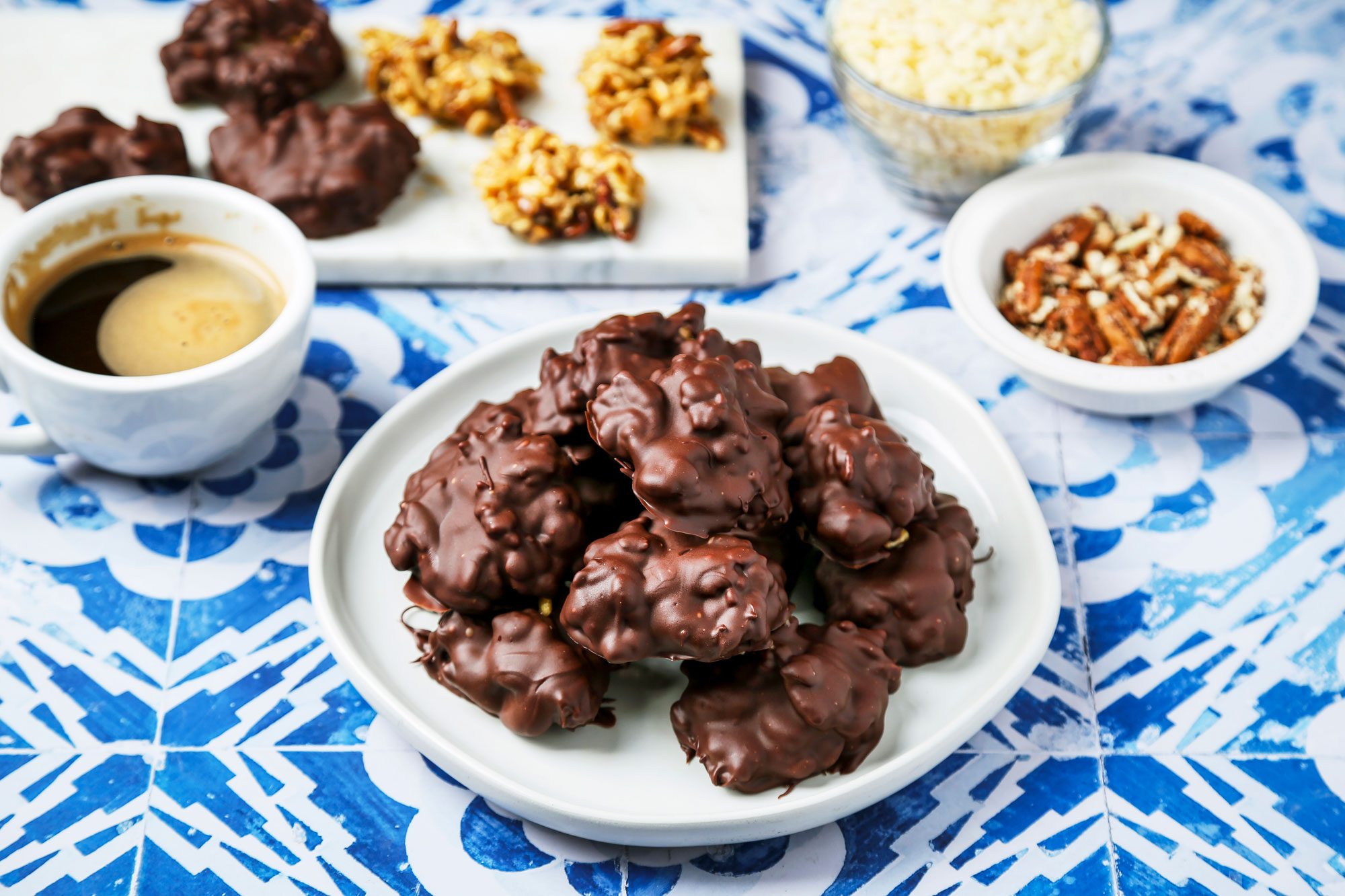 3/4th shot of Chocolate Billionaires; it is presented on a white plate and consists of several large, round chocolate coated treats; nearby, there is a small white bowl filled with chopped pecans as well as another bowl containing rice cereal; to the side, a cup of black coffee, the background features a decorative blue and white patterned surface