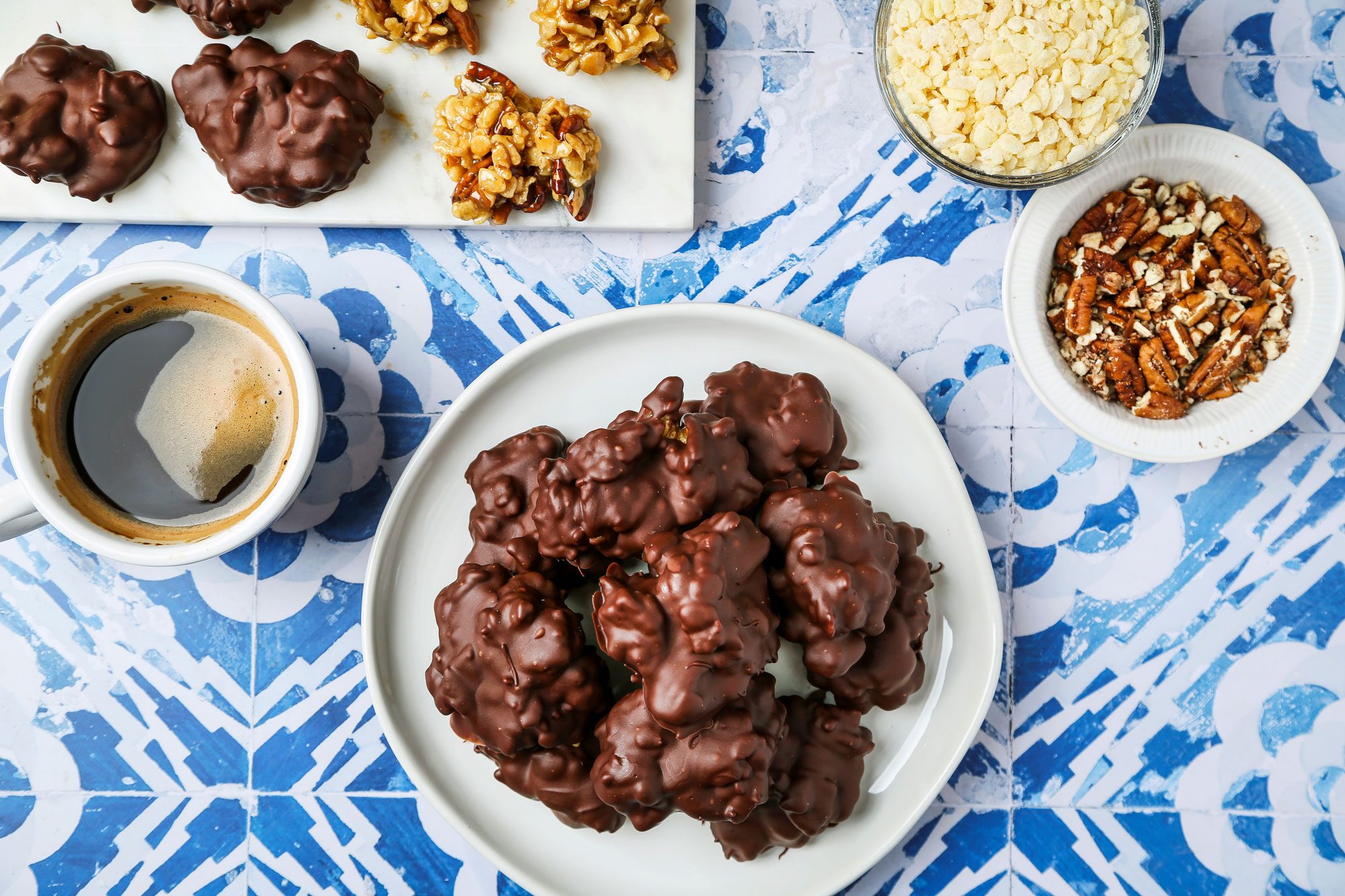overhead shot of Chocolate Billionaires; it is presented on a white plate and consists of several large, round chocolate coated treats; nearby, there is a small white bowl filled with chopped pecans as well as another bowl containing rice cereal; to the side, a cup of black coffee, the background features a decorative blue and white patterned surface