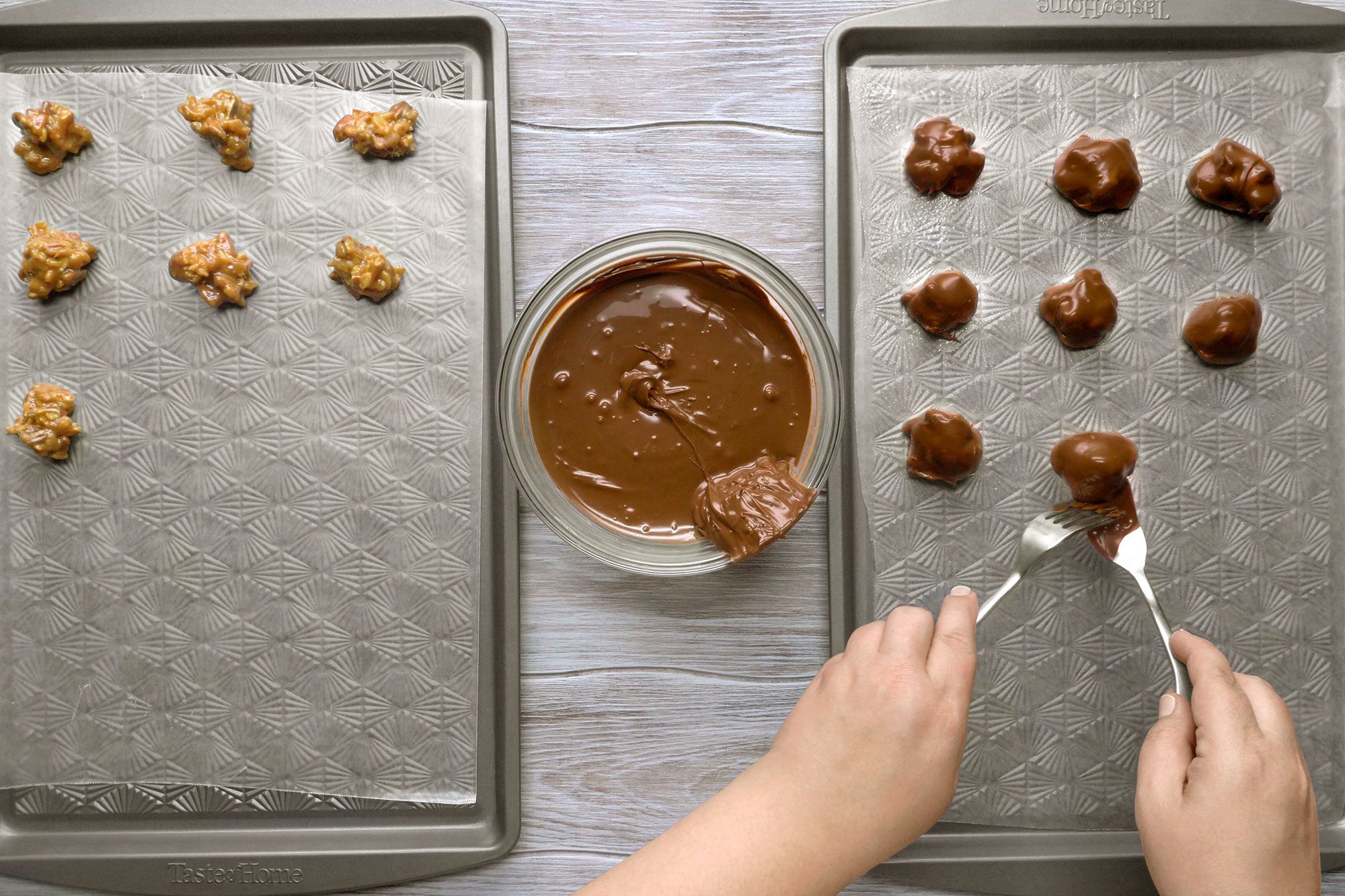 overhead shot of a person dipping peanut butter clusters into melted chocolate to make peanut butter cups; two baking sheets are on a wood table; one baking sheet holds the peanut butter clusters and the other holds the peanut butter clusters being dipped into chocolate; the person is holding two forks and is dipping a peanut butter cluster into the melted chocolate; a bowl of melted chocolate is near the baking sheets