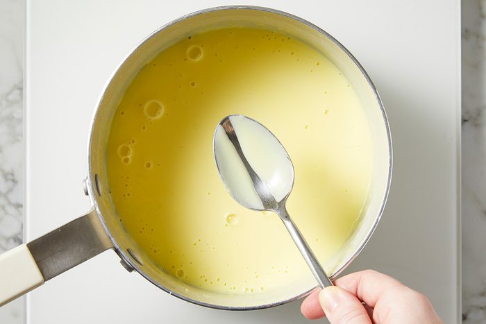 A hand holds a spoon over a saucepan containing smooth, creamy yellow sauce, possibly custard, on a light-colored surface.