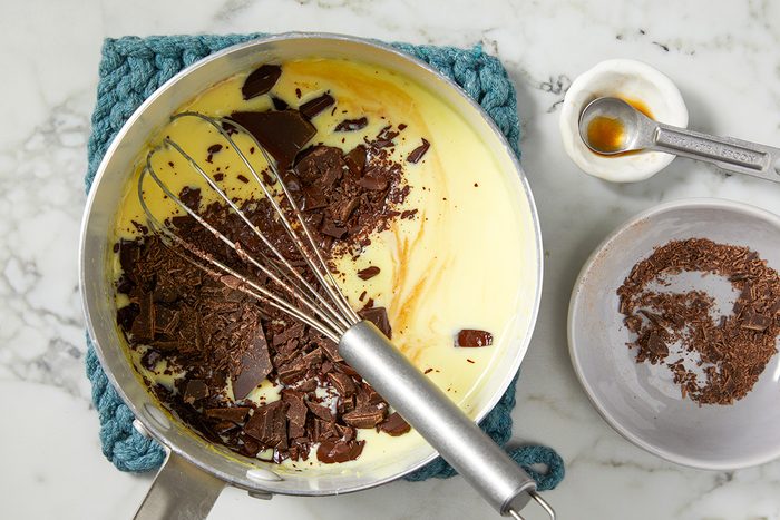 A saucepan on a blue trivet contains a yellow batter and chunks of dark chocolate, with a whisk resting inside. Nearby are a small bowl with chocolate shavings and a small cup of vanilla extract on a white marble surface.