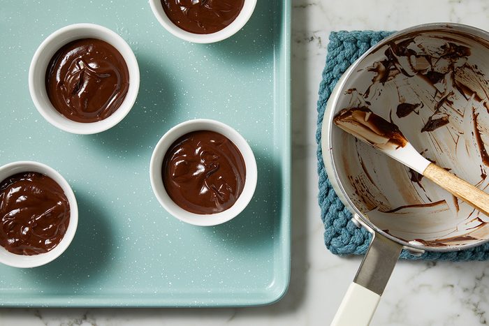 Four bowls of chocolate pudding are on a teal tray. Next to them is a saucepan with leftover chocolate residue and a wooden spatula, placed on a blue knitted mat on a marble surface.