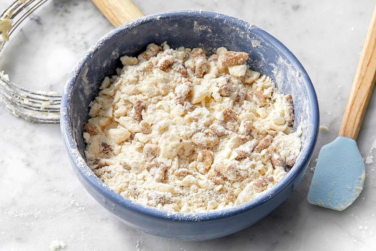 A blue bowl filled with crumbly dough mixture, featuring bits of butter and flour with nuts or raisins, sits on a marble counter. A pastry cutter and a blue spatula are nearby, suggesting preparation for baking.