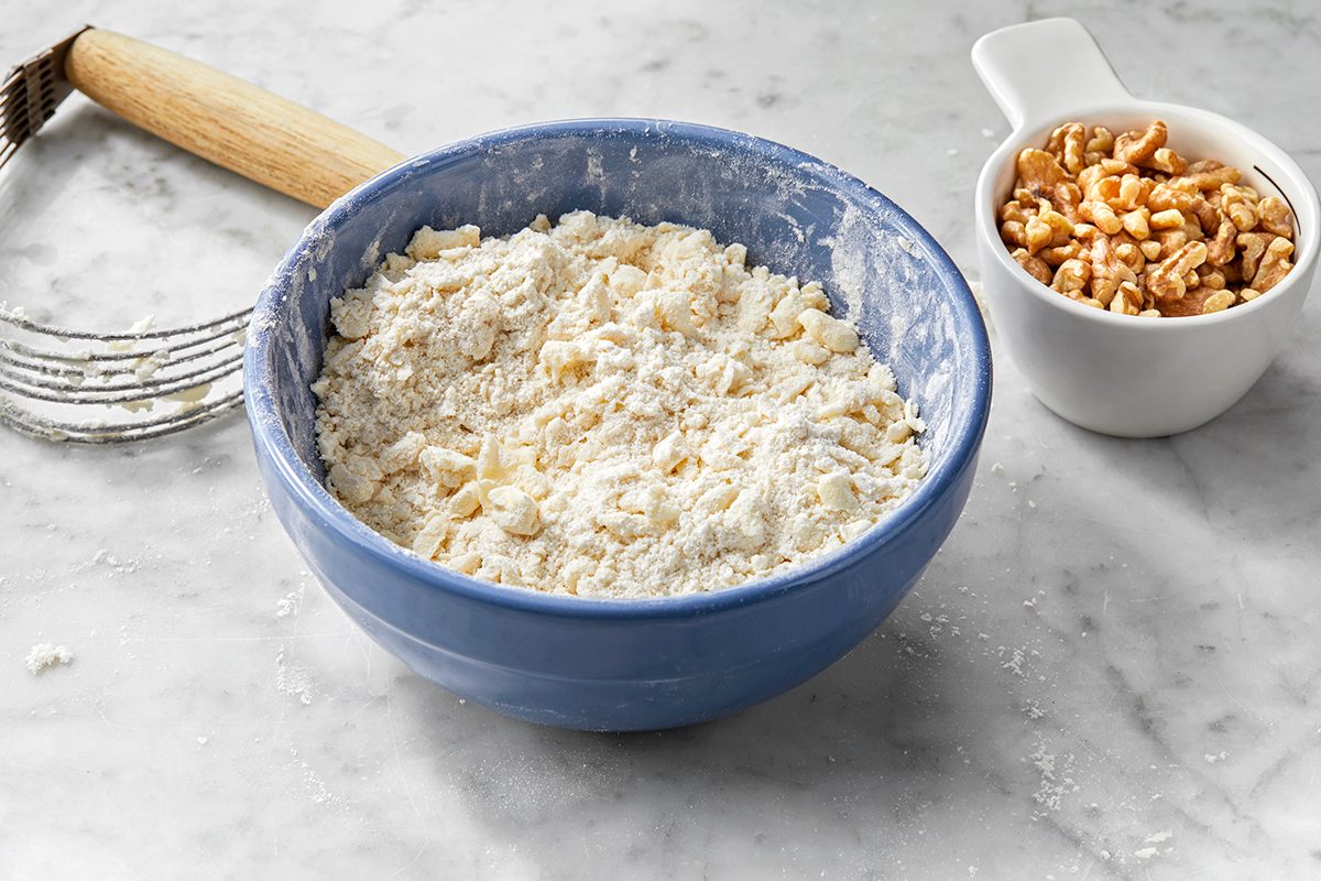 A blue bowl filled with a flour and butter mixture on a marble countertop. Beside it, a small white bowl holds chopped walnuts. A pastry cutter with a wooden handle lies nearby.