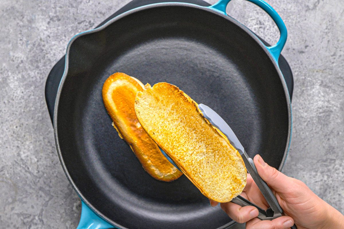 overhead shot of two slices of golden-brown bread being held by a pair of silver tongs over a black pan with a light blue handle, the bread appears to be toasted or grilled on one side, placed on a textured grey surface, likely a stovetop or kitchen counter