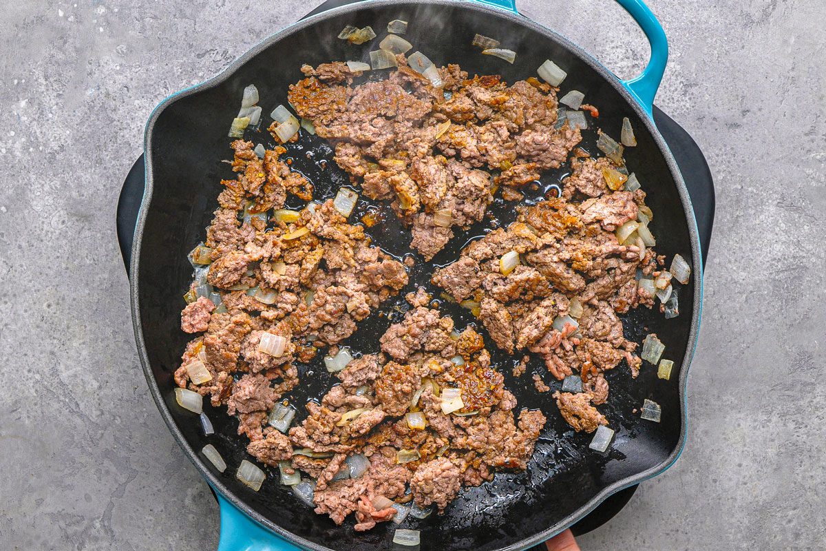 overhead shot of ground meat, cooking in a black pan, it is mixed with translucent diced onions, the pan is placed on a textured grey surface