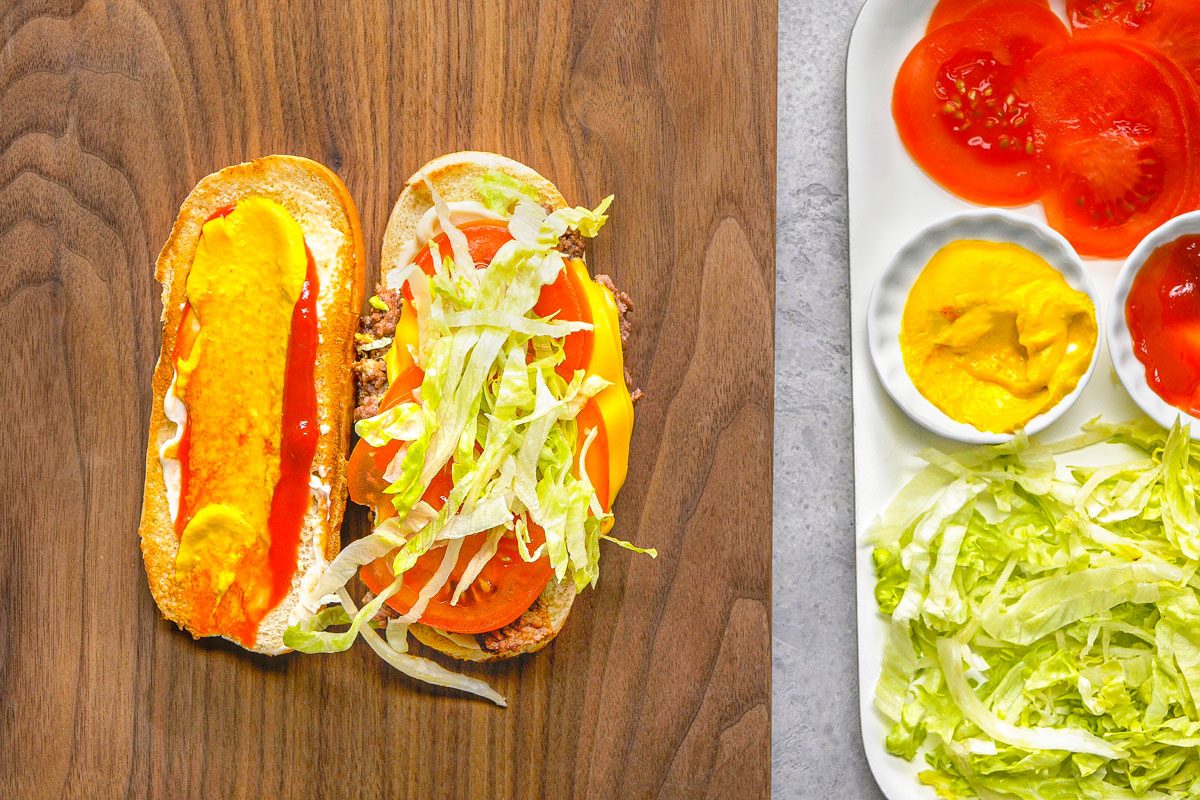 overhead shot of a partially assembled sandwich, on a wooden cutting board, the sandwich is made on a hoagie-style bun that is split open, one half is dressed with yellow mustard and red ketchup, while the other half contains a meat patty, melted cheese, and sliced tomatoes, topped with shredded lettuce, to the right, a white rectangular plate holds additional ingredients