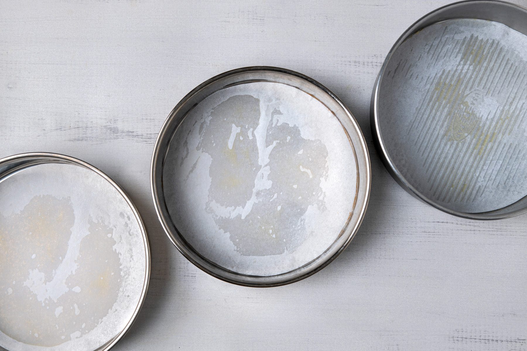 overhead shot of three round baking pans placed on a light wooden surface; all three pans are lined with parchment paper