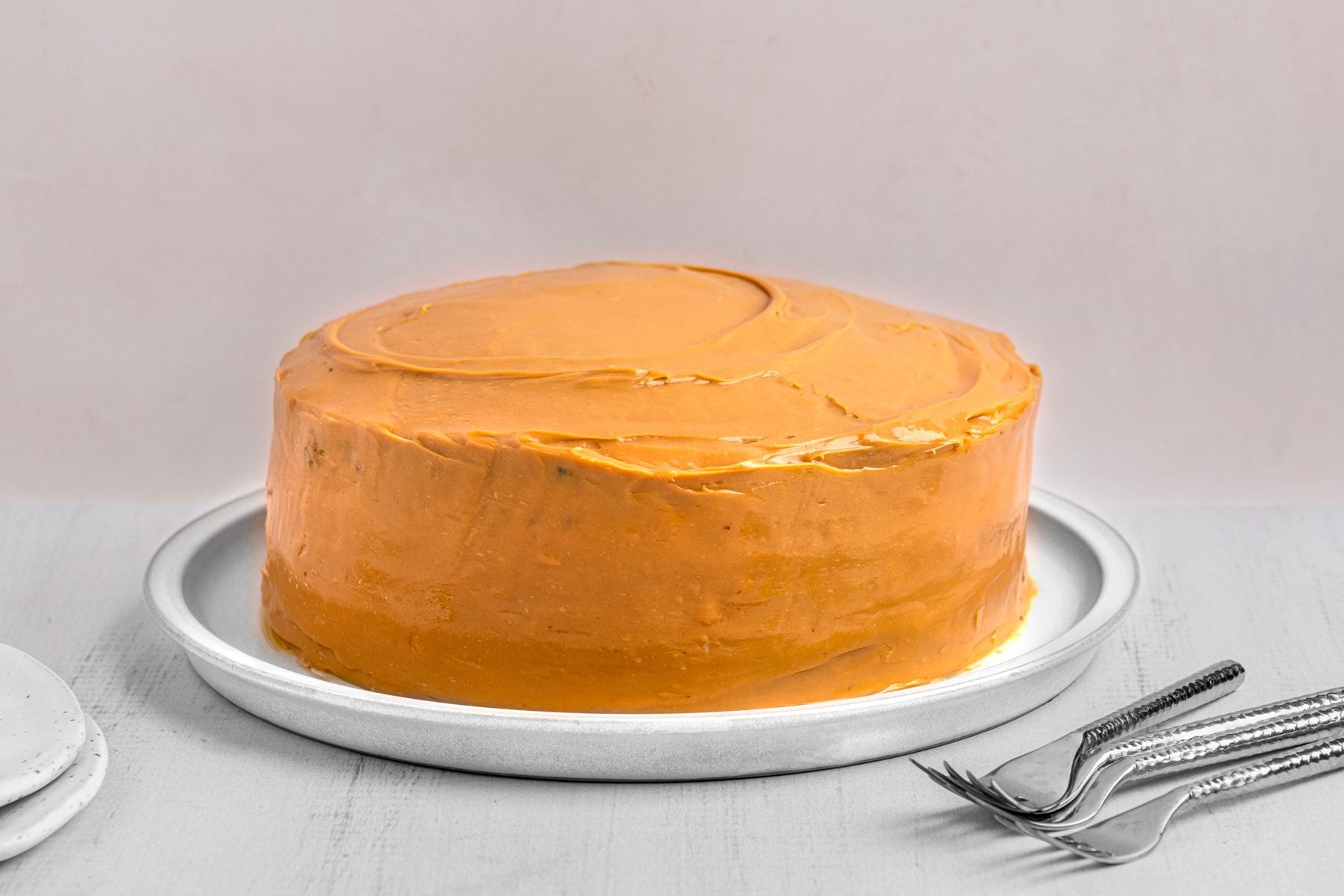 3/4th shot of a Dulce de Leche Cake placed on a round silver platter; in the background, there are a couple of white ceramic plates stacked together and a set of forks
