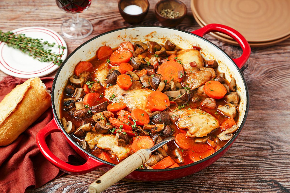 A red pot filled with a hearty stew featuring cooked chicken, sliced carrots, mushrooms, and herbs in a rich sauce. A piece of bread, a sprig of fresh thyme, and a glass of red wine are beside the pot on a wooden table.