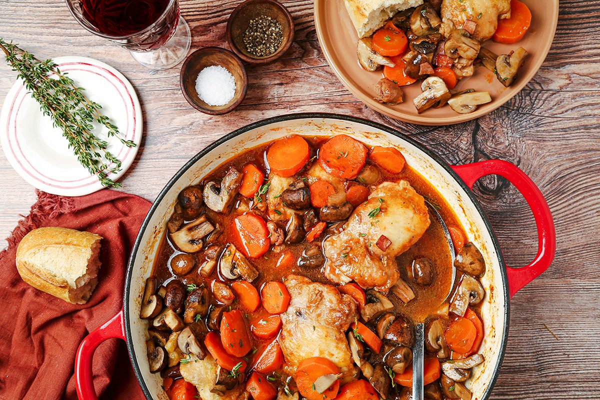 A rustic meal of chicken thighs, carrots, and mushrooms cooked in a red casserole dish. A plate with a serving sits nearby, surrounded by a glass of red wine, thyme sprigs, salt, pepper, and a bread roll on a wooden table.