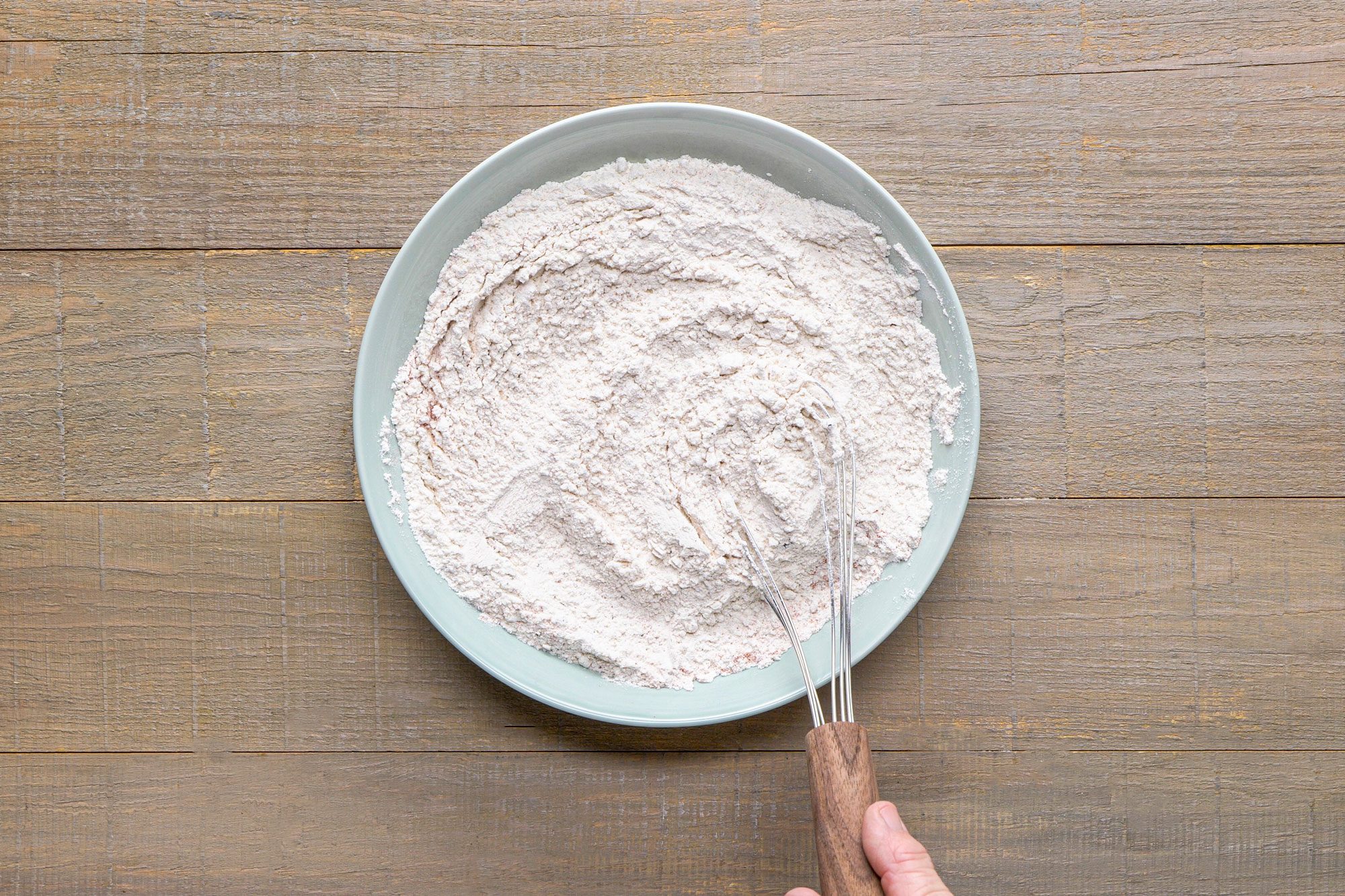 overhead shot of a large bowl filled with flour, a whisk, held by a hand, is positioned at the edge of the bowl, the background is a wooden surface with a subtle texture
