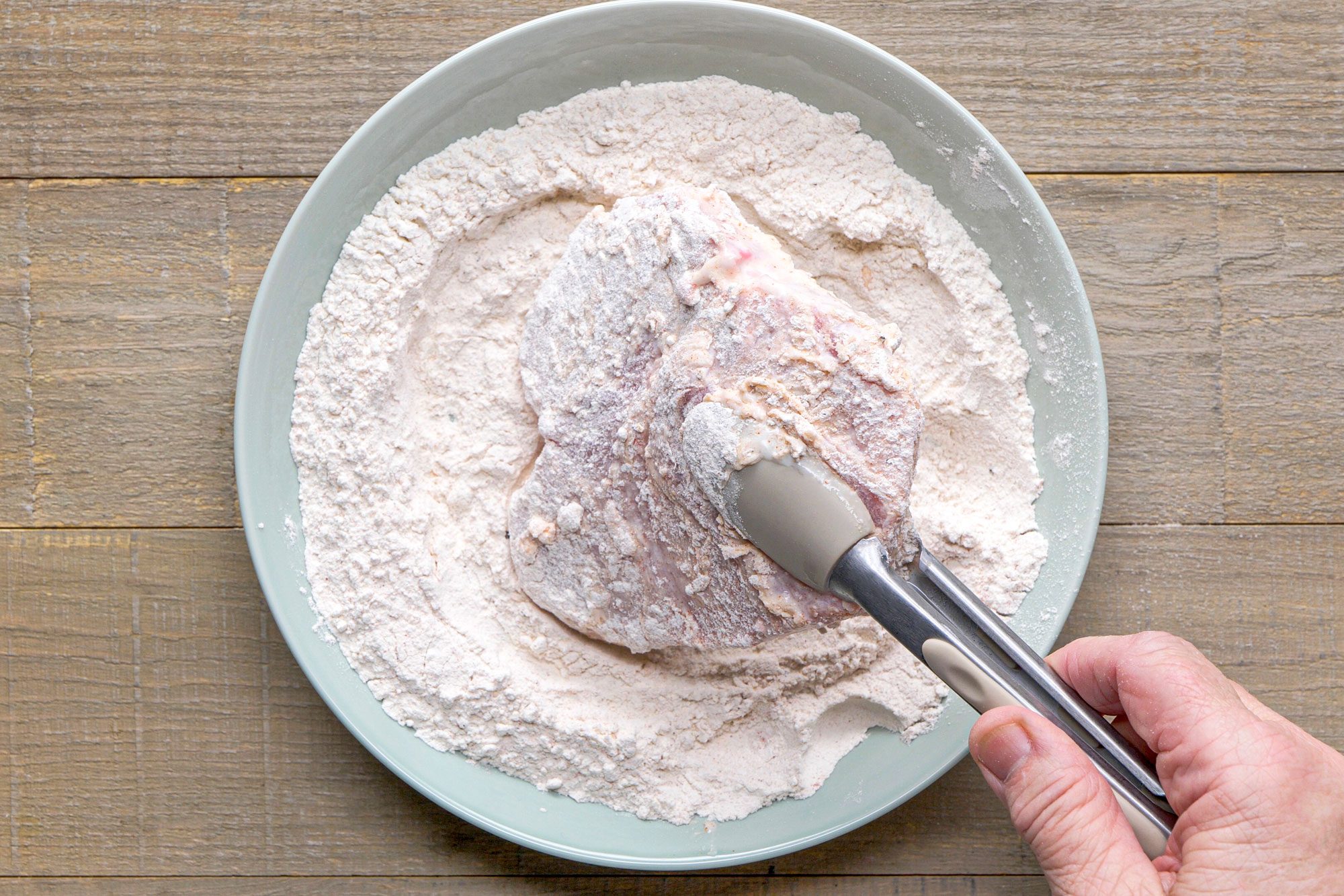 overhead shot of a hand holding a pair of tongs while coating a piece of meat in a flour mixture; the meat appears to be partially submerged in the flour, which is placed in a shallow, round dish; the background consists of wooden surfaces