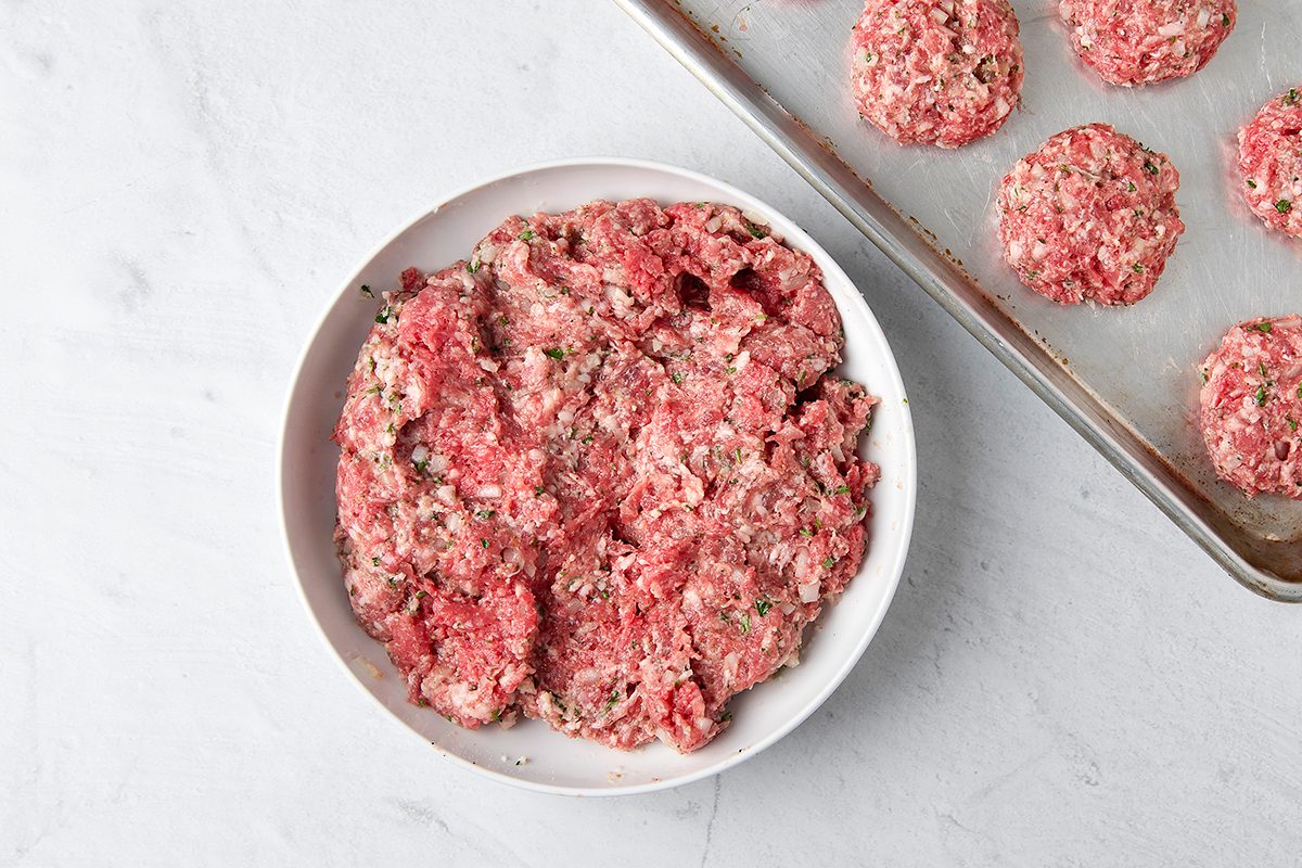 A bowl of raw ground meat mixed with herbs is on a white surface. Next to it, a baking sheet displays several formed meatballs.