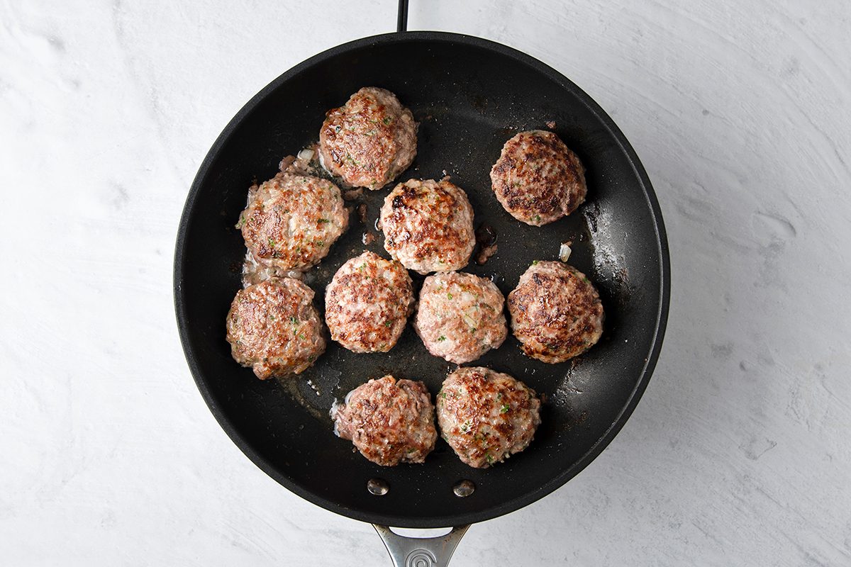 A non-stick skillet with nine cooked round meat patties, slightly browned, arranged on a light gray countertop.
