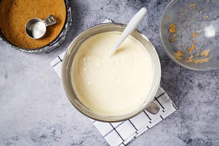 A bowl filled with creamy batter and a spatula sits on a checkered cloth. Nearby, there's a round pan with crushed biscuits and a small measuring cup.