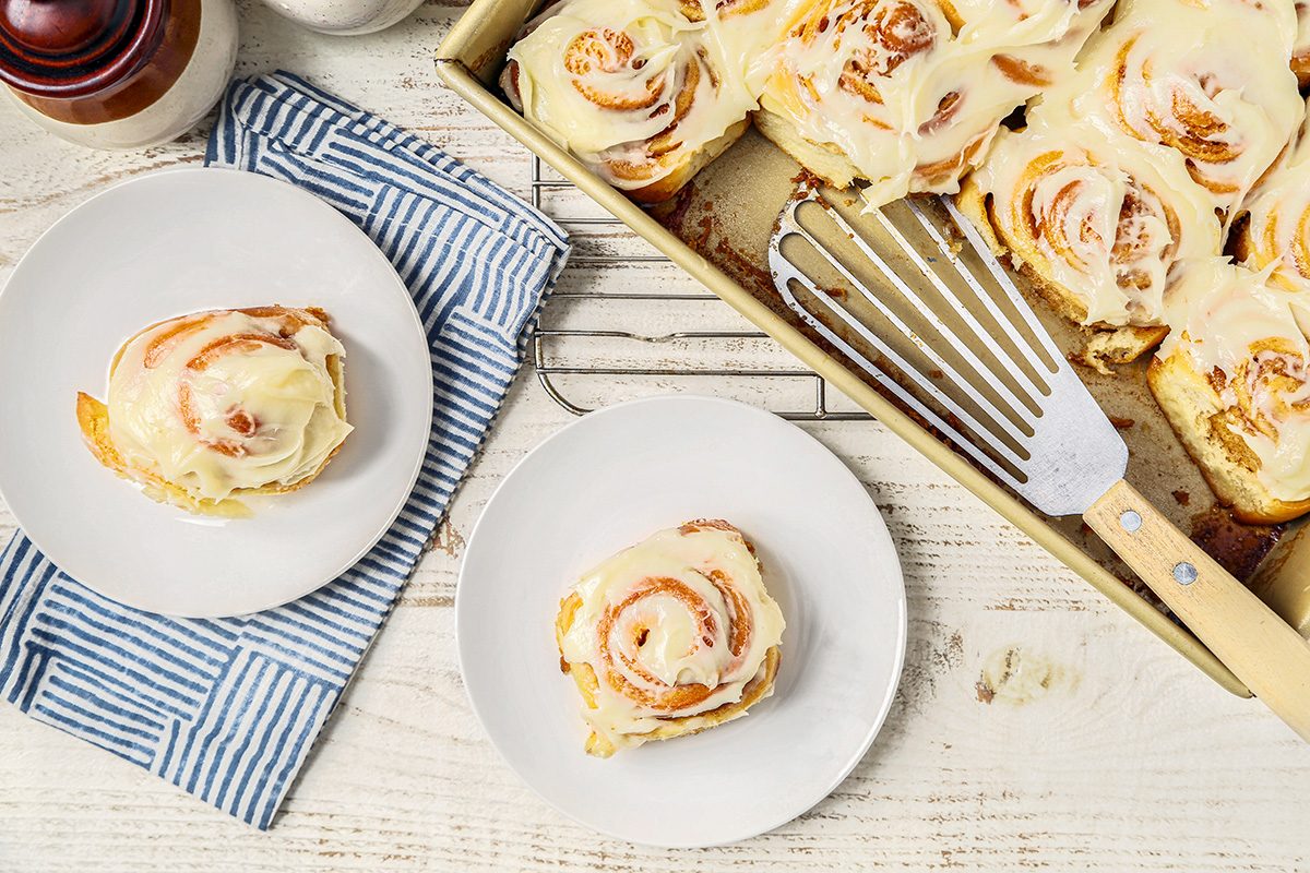 Cinnamon rolls with icing on a white wooden table. Two rolls are on separate plates, beside a pan of more rolls. A striped cloth and a metal spatula are nearby.