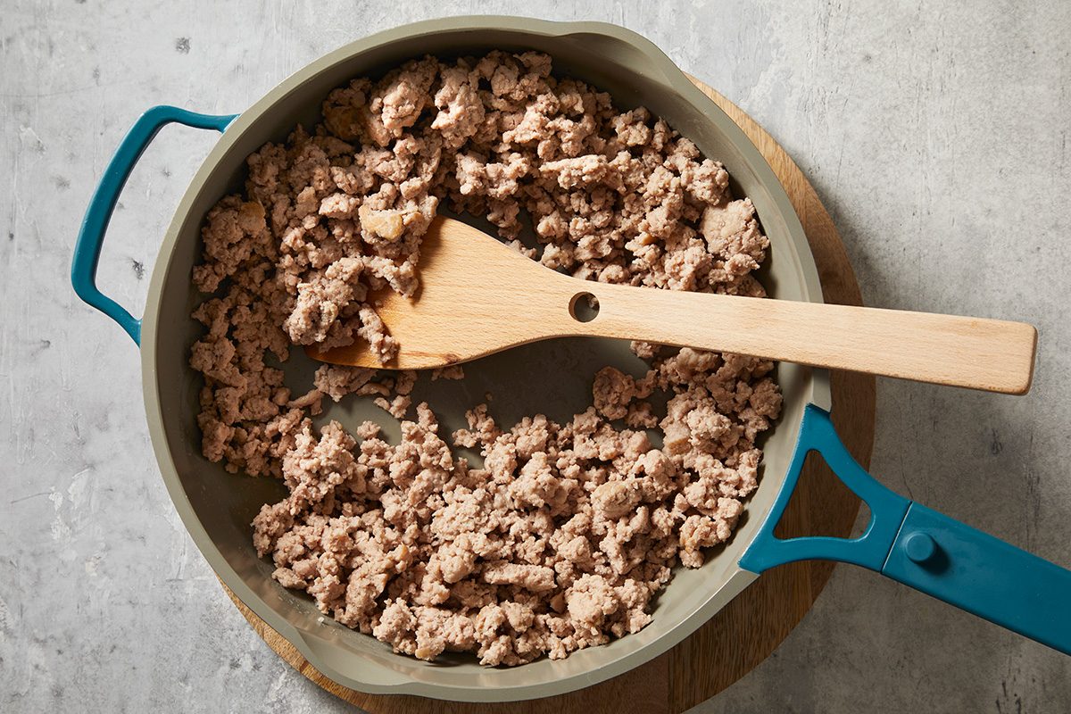 overhead shot of a large, shallow pan filled with cooked, crumbled meat, of ground turkey, A wooden spoon rests in the mixture