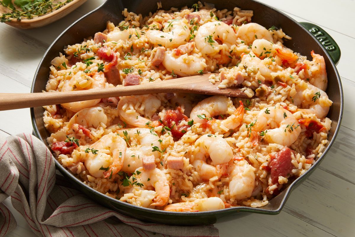 3/4th shot of Ham and Shrimp Jambalaya in a deep skillet; a wooden spoon rests atop the mixture, the surrounding area includes a patterned towel