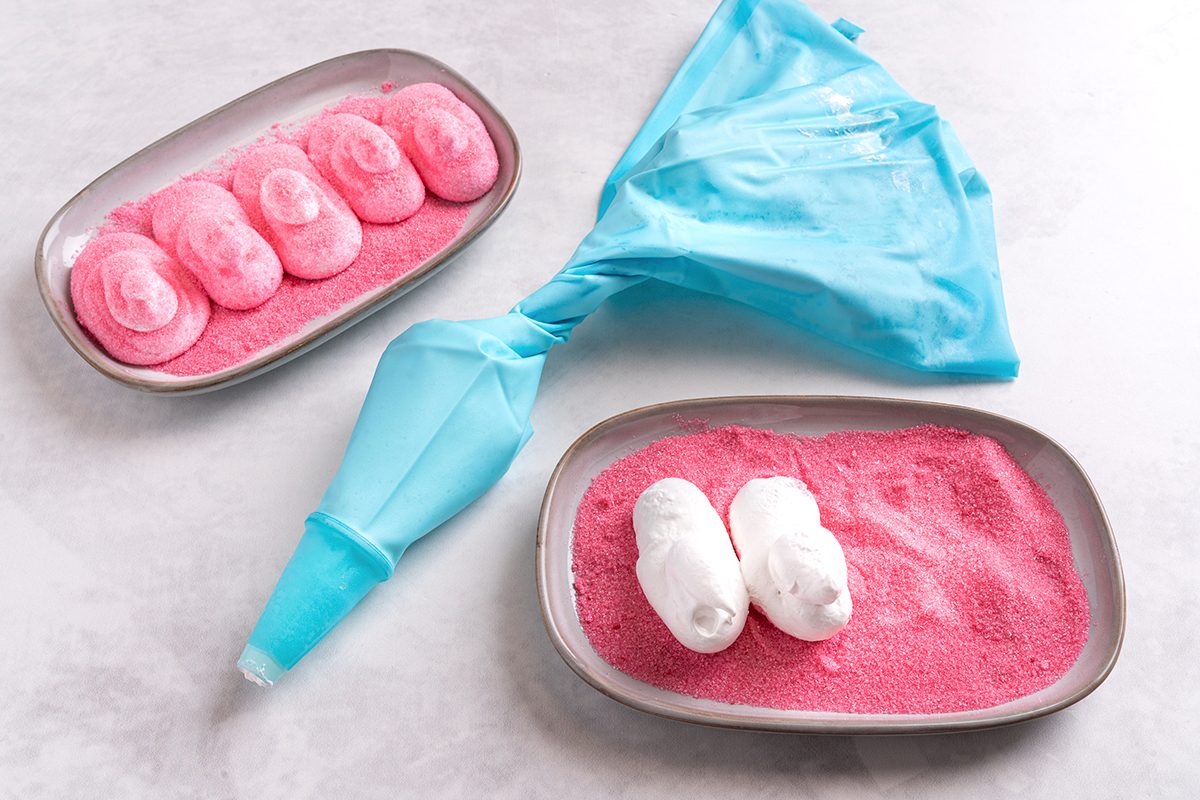 A piping bag with light blue icing lies on a surface. To its left, a tray holds five pink icing shapes on pink sugar. To its right, another tray contains two white icing shapes on pink sugar.
