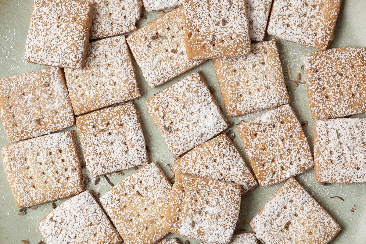 Square cookies dusted with powdered sugar are arranged in a scattered pattern on a light green surface. The cookies have a golden-brown color and a textured appearance.