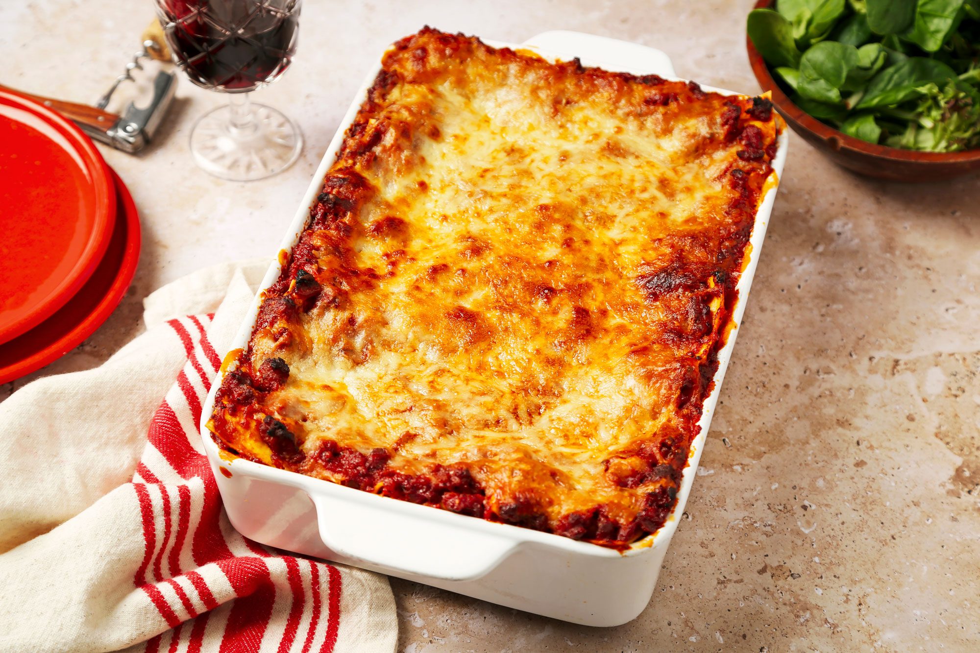3/4th shot of a freshly baked lasagna in a white rectangular dish; the dish is placed on a light colored countertop, accompanied by a red striped linen towel and two red plates to the side; in the background, there is a glass of dark red wine and a bowl of fresh greens, likely spinach