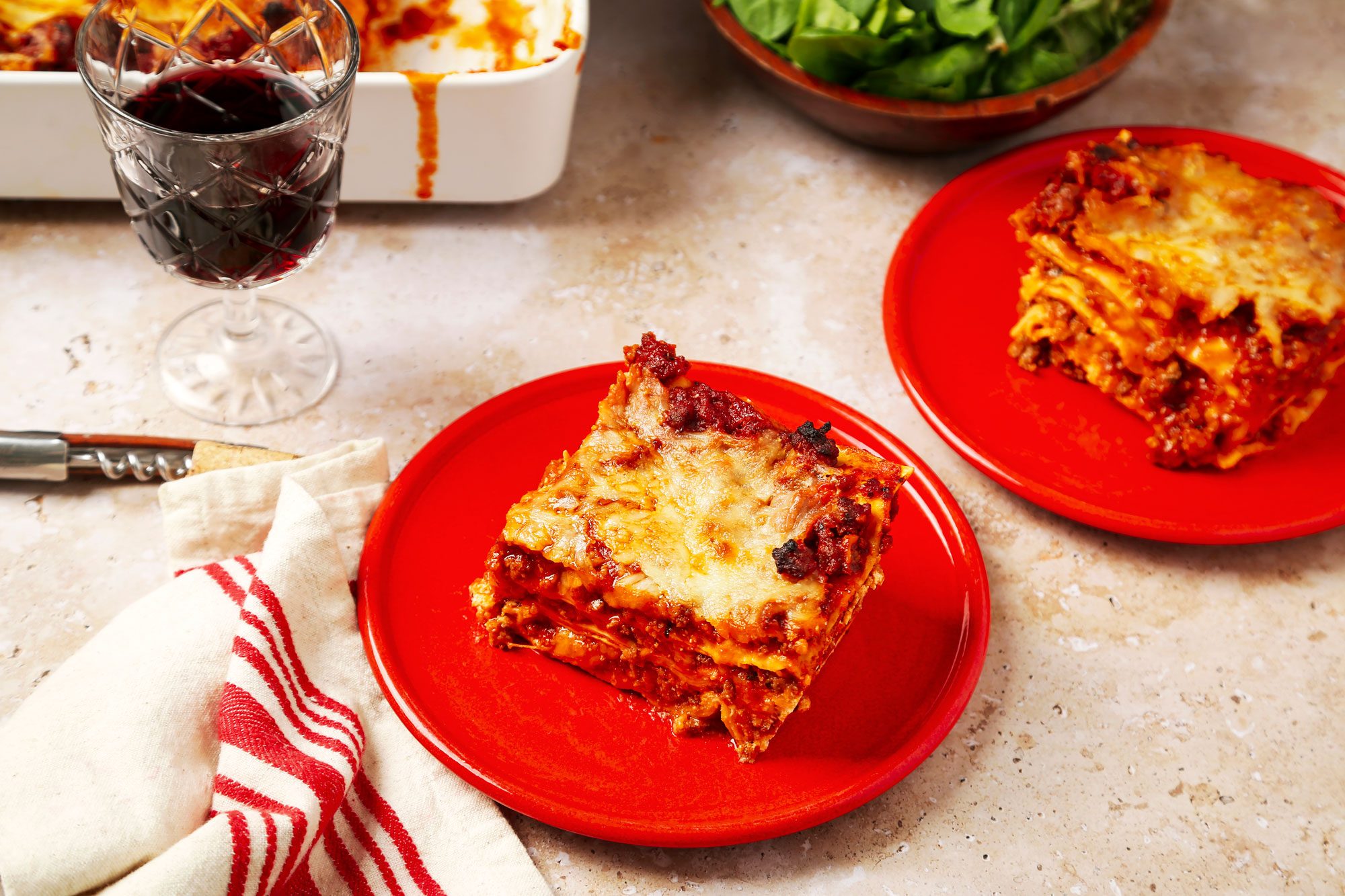 3/4th shot of a lasagna plated on a red dish; two pieces of lasagna are visible, one prominently in the foreground; there is also a second piece in the background on a similar plate, beside the lasagna, a clear wine glass filled with dark red wine sits on the table, to the side, a white cloth napkin with red stripes is folded neatly, and a silver corkscrew can be seen resting nearby; in the background, there's a bowl of fresh green spinach leaves