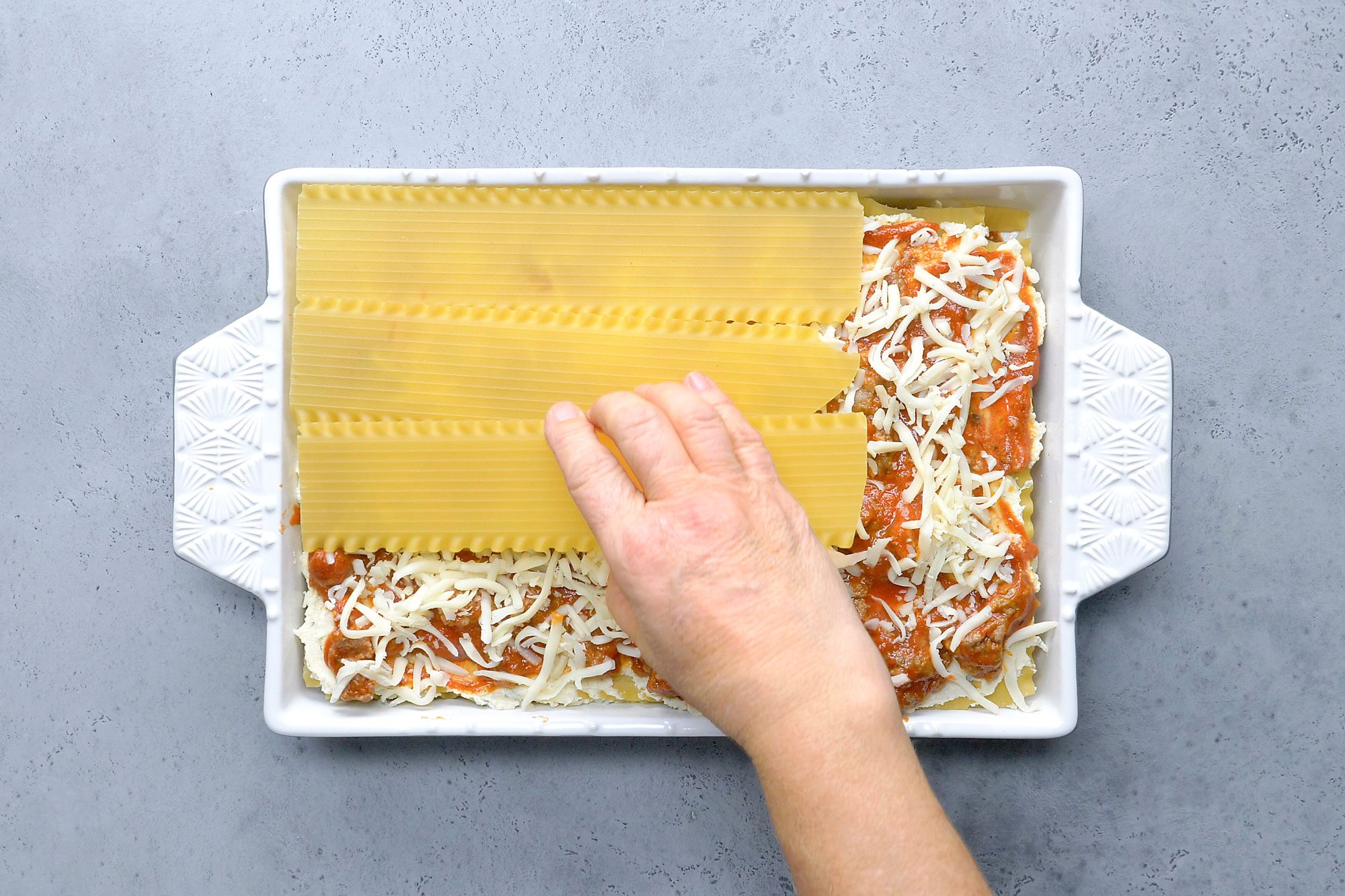 Overhead shot of spread 1 cup meat sauce into a greased baking dish; Layer with 4 noodles; half of the ricotta cheese mixture; 1 cup meat sauce and 1 cup mozzarella cheese; Repeat layers; grey surface;