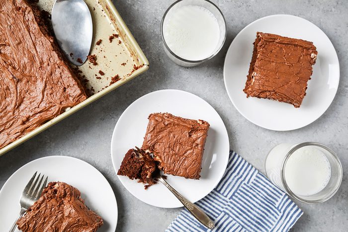 Top view of three plates with chocolate brownies, each beside a glass of milk on a gray surface. A baking tray with remaining brownies and a serving spatula is on the left. A blue and white striped napkin is placed near the plates.