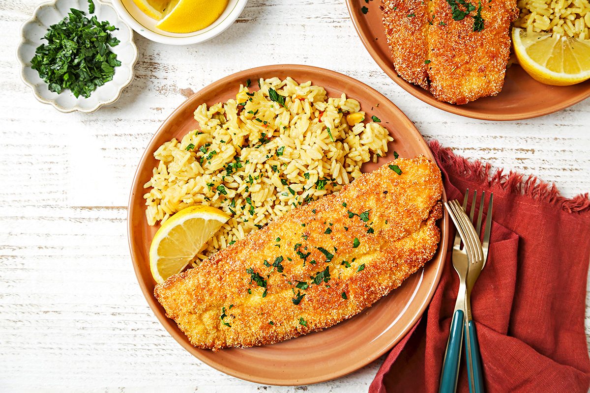 A plate with breaded fish fillet topped with chopped parsley, rice pilaf, and a lemon wedge on a light wooden surface. Next to it are a small dish of parsley, a red cloth napkin, and a fork and knife. A second plate is partially visible.