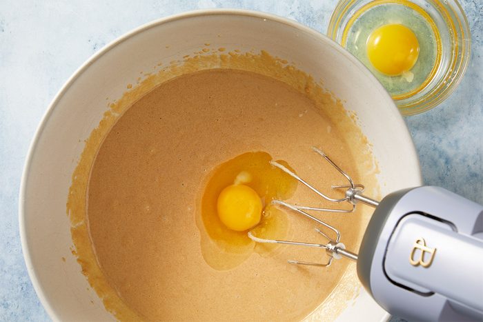 overhead shot of a large mixing bowl filled with a batter; at the center of the batter, there is a raw egg with its yolk visible, beside the bowl, a handheld mixer is positioned, its beaters resting in the batter; a smaller bowl containing another whole egg sits in the background
