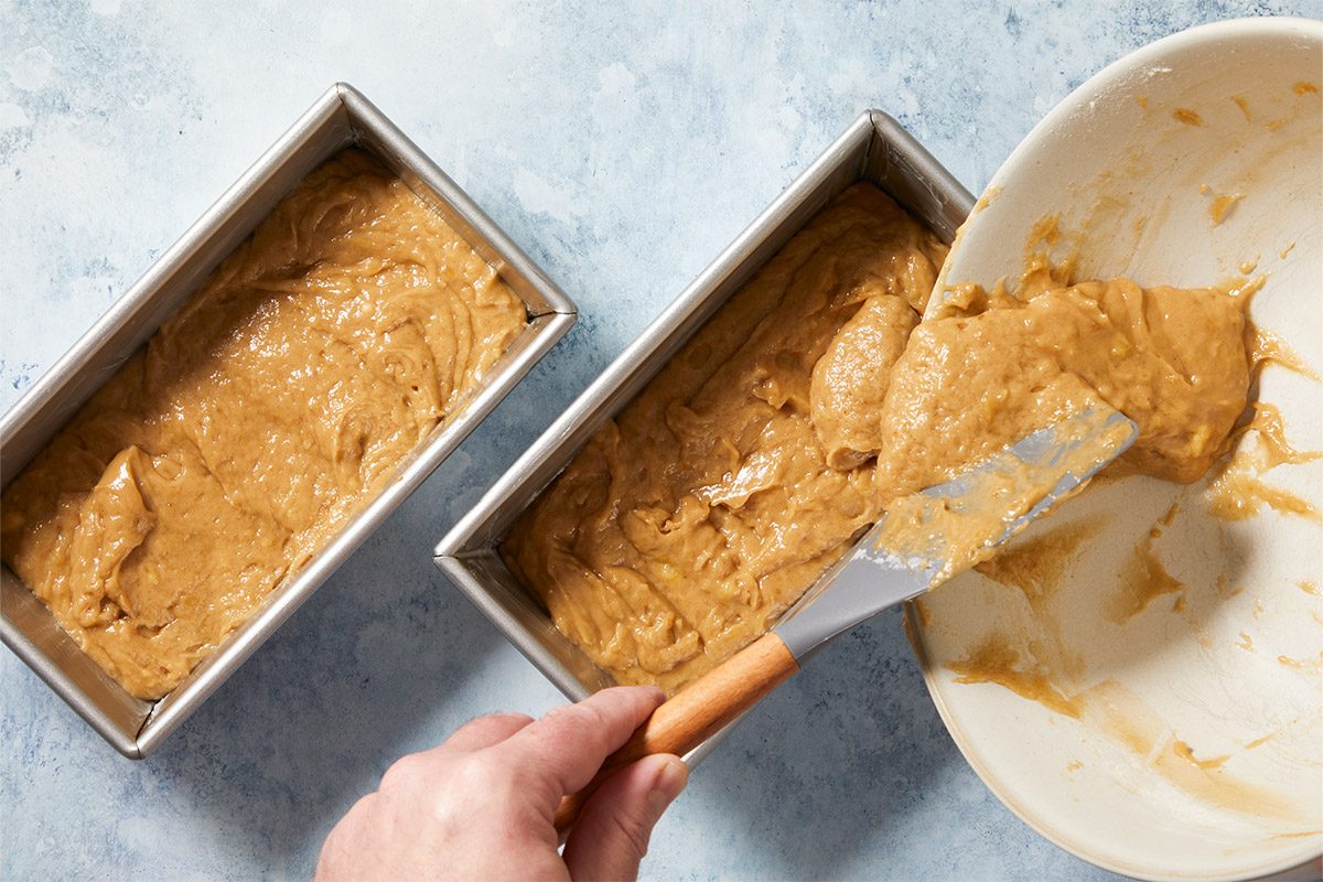 overhead shot of two metallic baking pans filled with a thick batter and a light brown color; a hand is holding a spatula, which is coated with more of the batter, while one of the pans appears slightly fuller than the other; the background has a subtle blue color