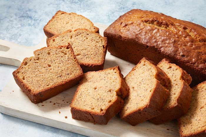 3/4th shot of Peanut Butter Banana Bread, with a golden brown crust; Slices of the bread are arranged on a wooden cutting board, The loaf is positioned upright, The background features a light, textured surface