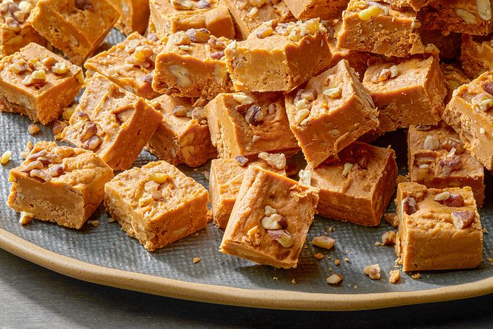 A plate filled with square, golden brown pieces of fudge topped with chopped nuts. The fudge pieces are closely packed and have a creamy texture, sprinkled with assorted nuts on top for a crunchy finish.