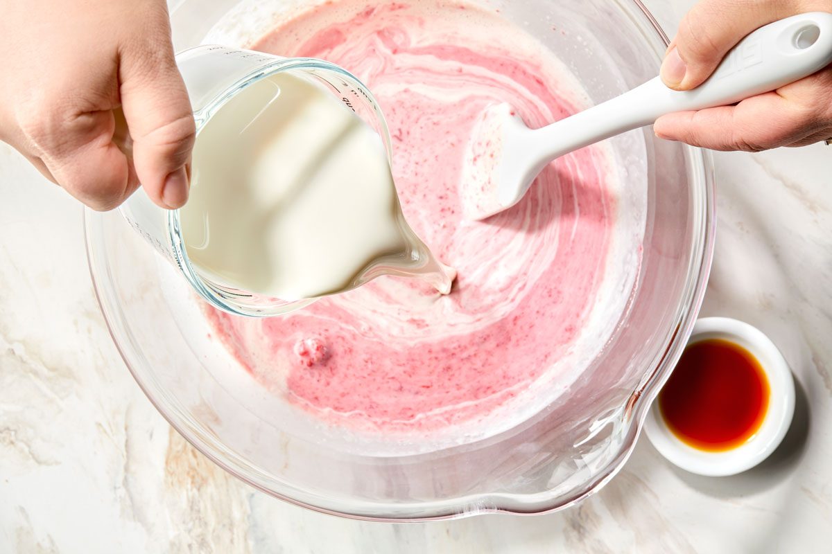 A person pouring whipping cream into a bowl of raspberries and sugar.
