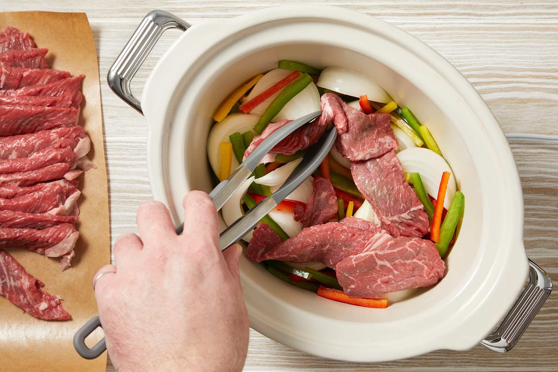Overhead shot of placing peppers and onion in a 5-qt. slow cooker. Top with beef. Kept in a white baking dish over a light wooden countertop.