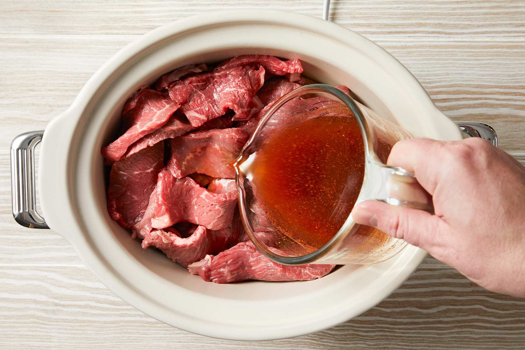 Overhead shot of pouring over the water, vinegar, lime juice and seasonings mixture over the meat in a white baking dish over a light wooden countertop