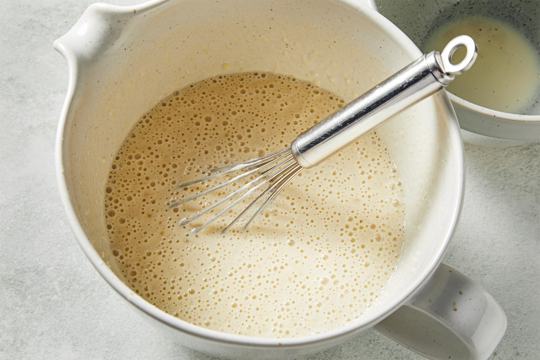 A steel whisk being used to whisk up a white and foamy liquid in a large white mixing bowl.