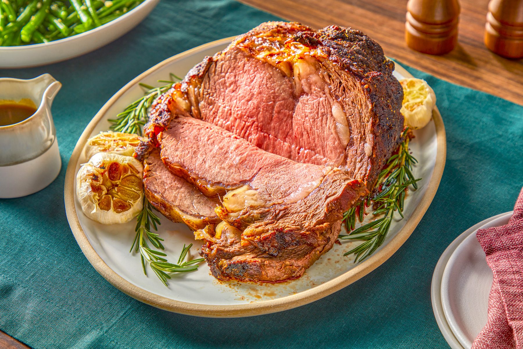 3/4 angle view shot of Standing Rib Roast; served on a large plate; with sauce and cutlery; mpty plates with a napkin over them; green tablecloth on a wooden surface;