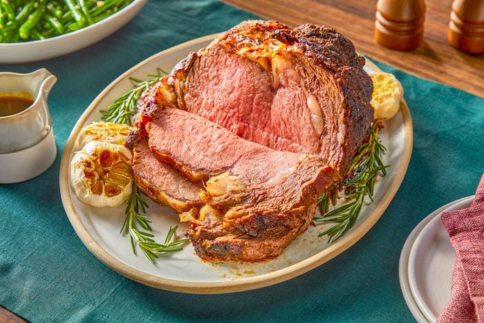 3/4 angle view shot of Standing Rib Roast; served on a large plate; with sauce and cutlery; mpty plates with a napkin over them; green tablecloth on a wooden surface;