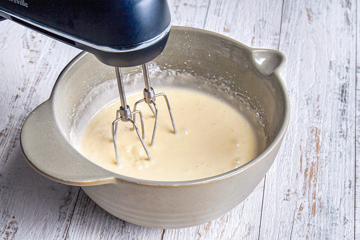 3/4th shot of a mixing bowl containing a creamy, thick batter or mixture; An electric hand mixer is positioned above the bowl, with its beaters immersed in the mixture; The surface beneath the bowl is made of white wood