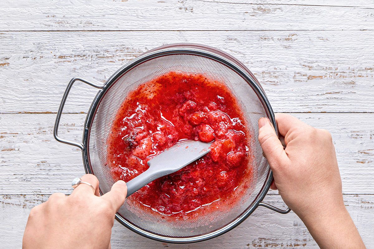 overhead shot of a person is straining a mixture of bright red raspberry puree through a fine mesh sieve; the sieve is positioned above a white wooden surface, one hand is holding the sieve while the other is using a spatula to press the puree, extracting the juice and leaving the seeds behind;