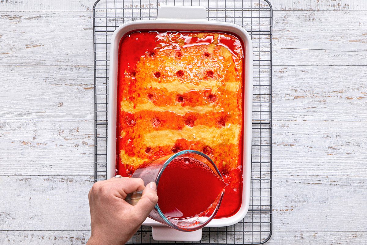 overhead shot of a rectangular cake with a light golden color topped with a red syrup that has pooled around it, the cake is sitting in a white baking dish placed on a wire rack above a wooden surface, a hand can be seen pouring bright red liquid from a measuring cup onto the cake