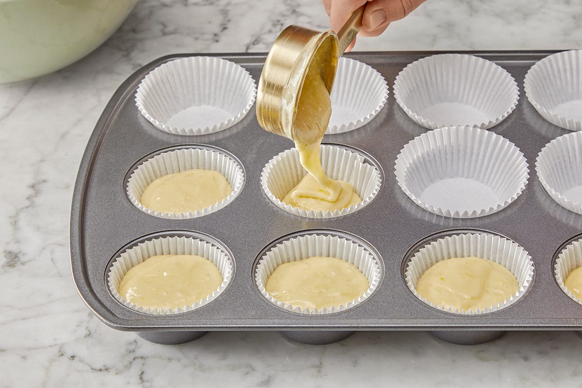 A hand pours cupcake batter into white paper liners in a muffin tin placed on a marble countertop.