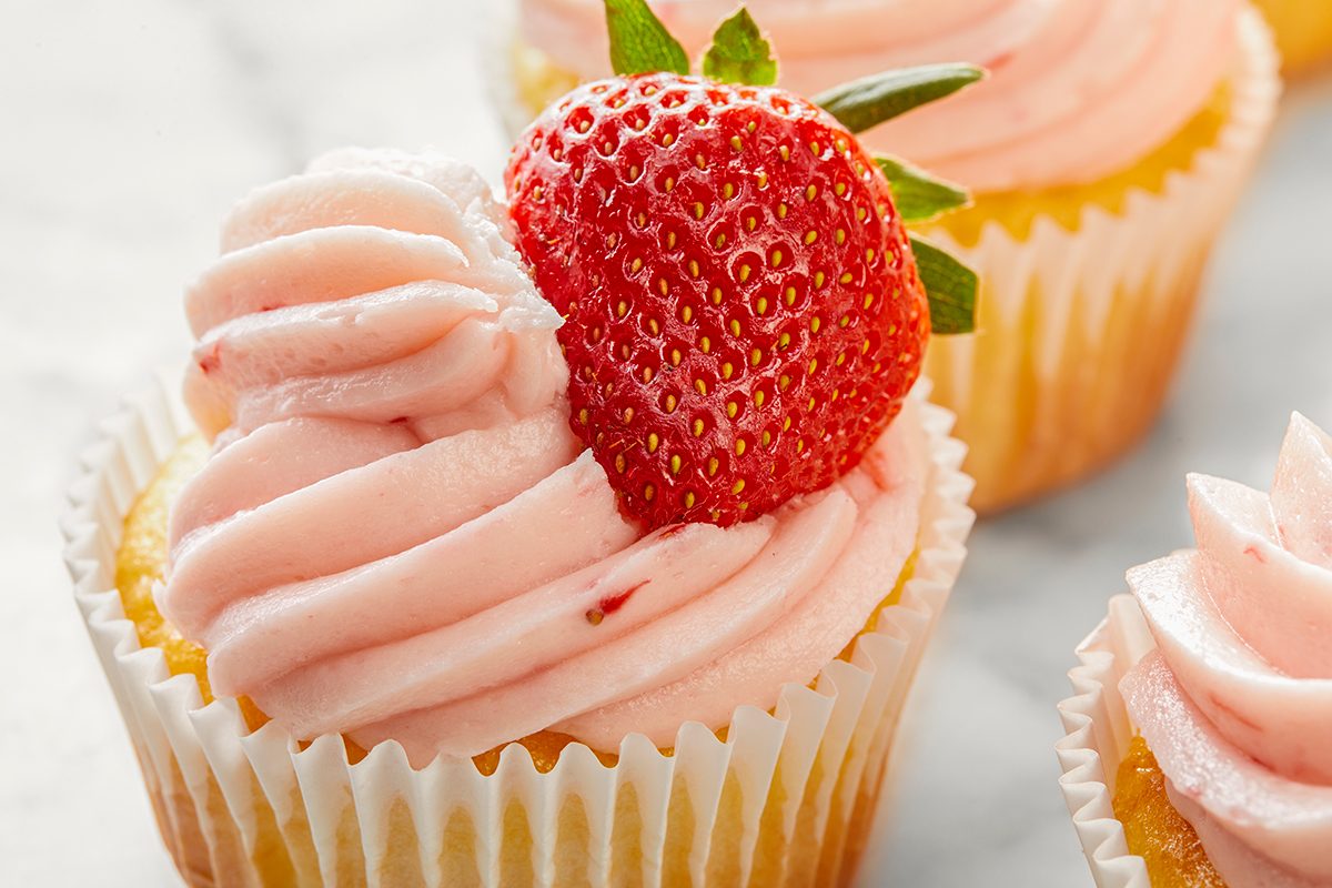 A close-up of a cupcake topped with pink strawberry frosting and a fresh strawberry slice. The cupcake rests on a marble surface, with another similar cupcake visible in the background.