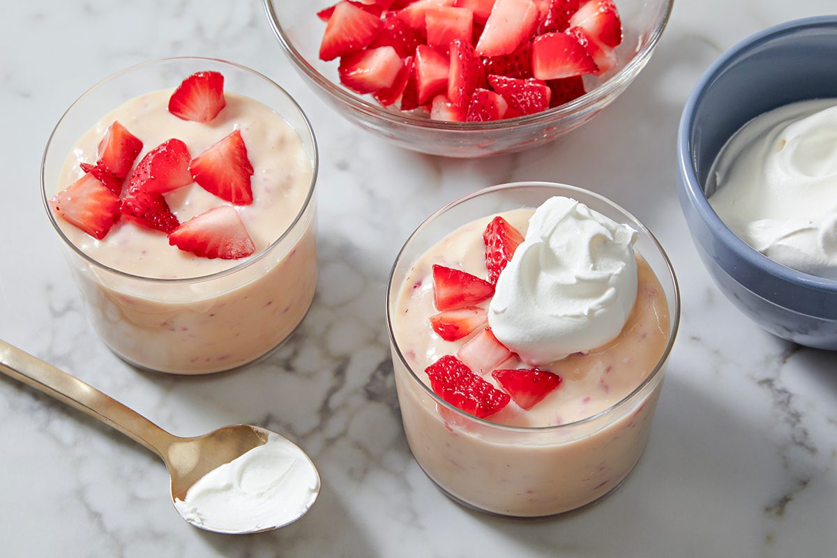 Two cups of strawberry mousse topped with sliced strawberries and whipped cream on a marble surface. A bowl of sliced strawberries and a bowl of whipped cream are in the background, alongside a spoon with whipped cream on it.