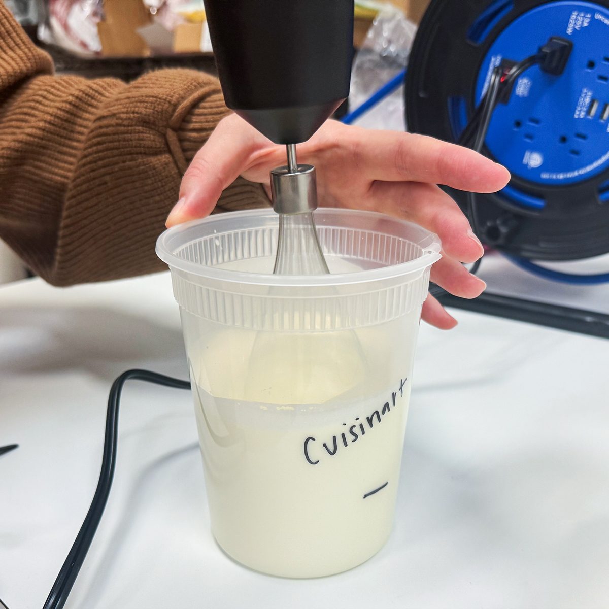 A person uses a handheld milk frother to mix liquid in a plastic cup labeled "Cuisinart" on a table. A blue object is visible in the background.
