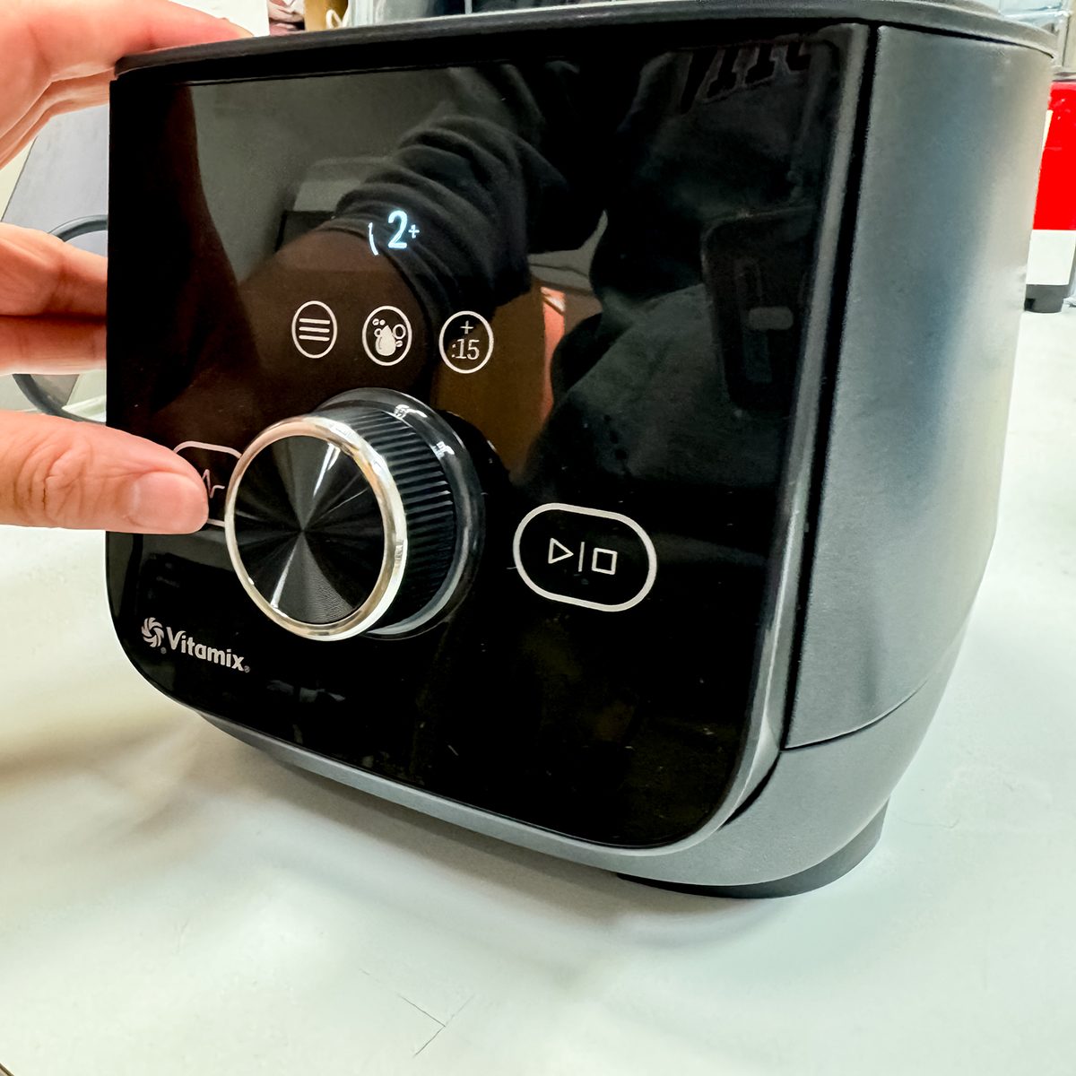 Close-up of a person adjusting the settings on a black Vitamix appliance. The front panel displays various function icons and a round central knob, with a digital screen above showing the number 2.